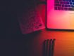 A laptop and a notebook with sketches on a wooden table, illuminated by colorful lighting