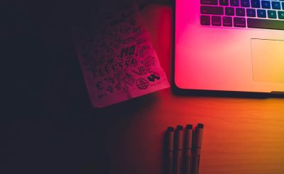 A laptop and a notebook with sketches on a wooden table, illuminated by colorful lighting
