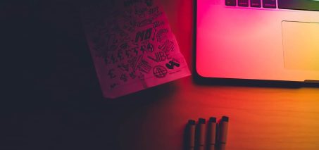 A laptop and a notebook with sketches on a wooden table, illuminated by colorful lighting