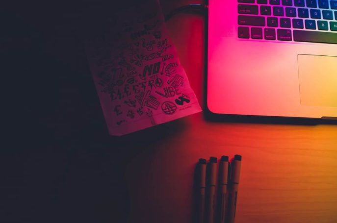 A laptop and a notebook with sketches on a wooden table, illuminated by colorful lighting